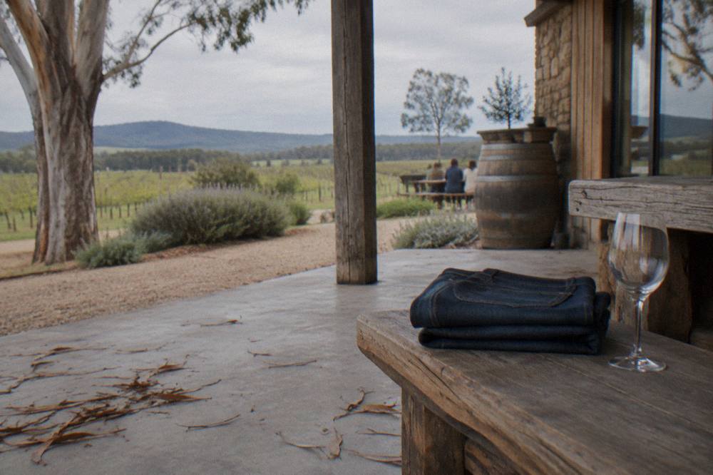 A pair of dark denim jeans folded on a rustic timber bench beside a wine glass.