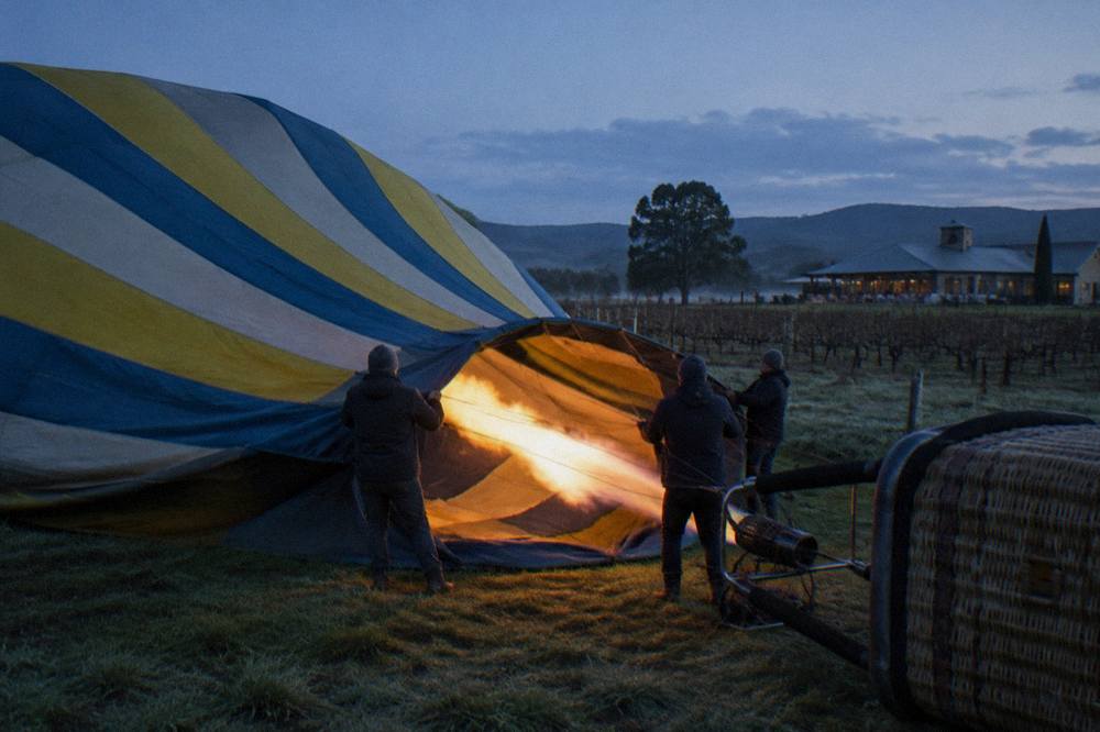 A striped hot air balloon being inflated on a paddock beside vineyards before dawn