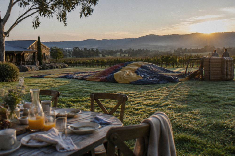 A set breakfast table on a lawn with vineyards and a deflated balloon in the background