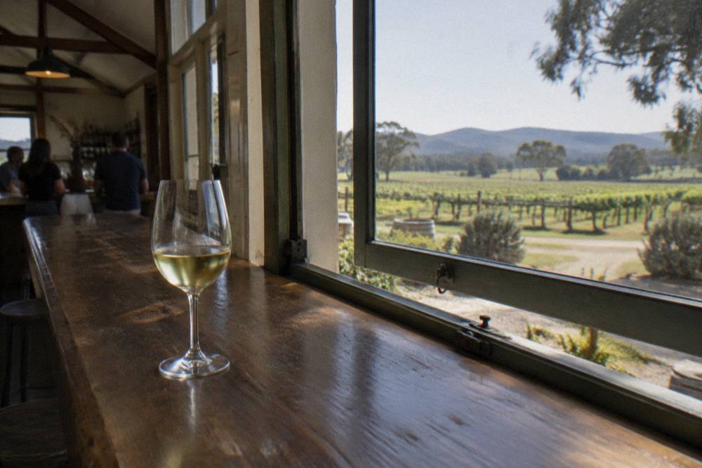 A glass of pale white wine on a timber tasting bar beside an open vineyard window