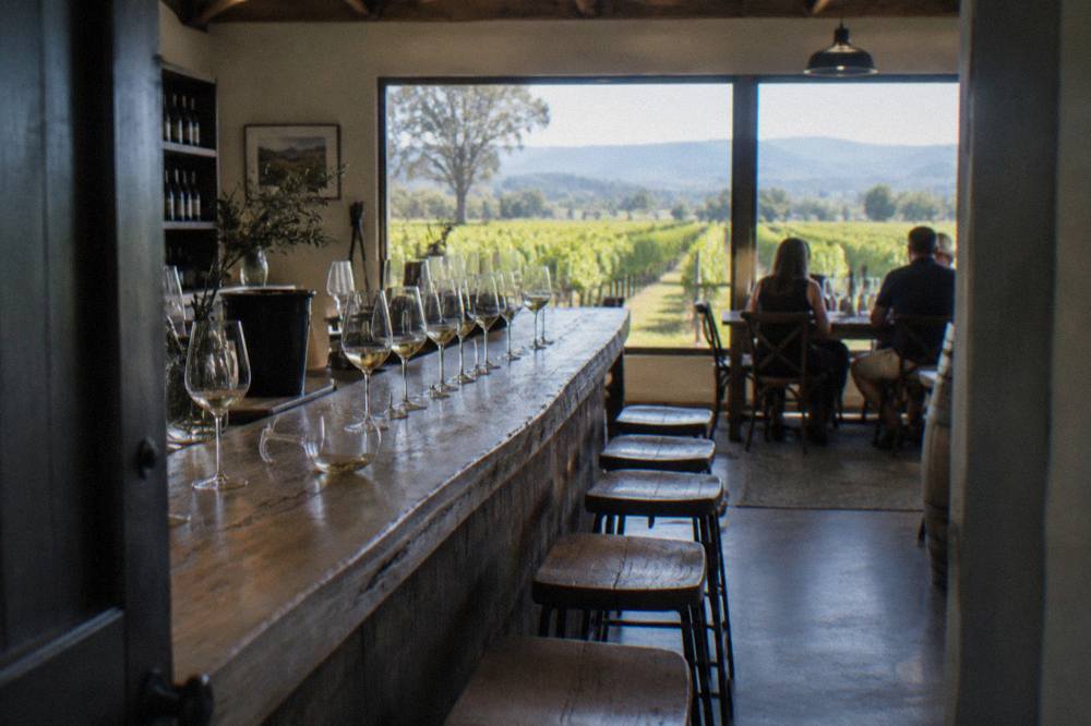 A row of white wine glasses lined along a timber tasting bar with vineyard rows visible through the window