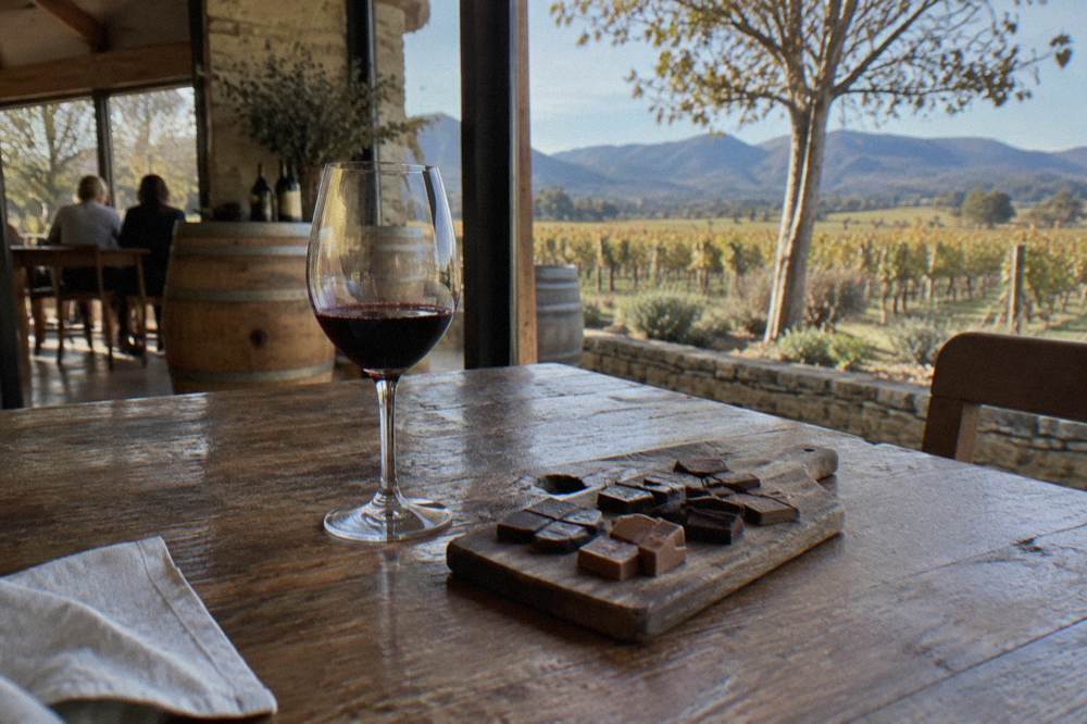 An artisan chocolate board with dark and milk chocolate squares beside a glass of red wine on a timber table