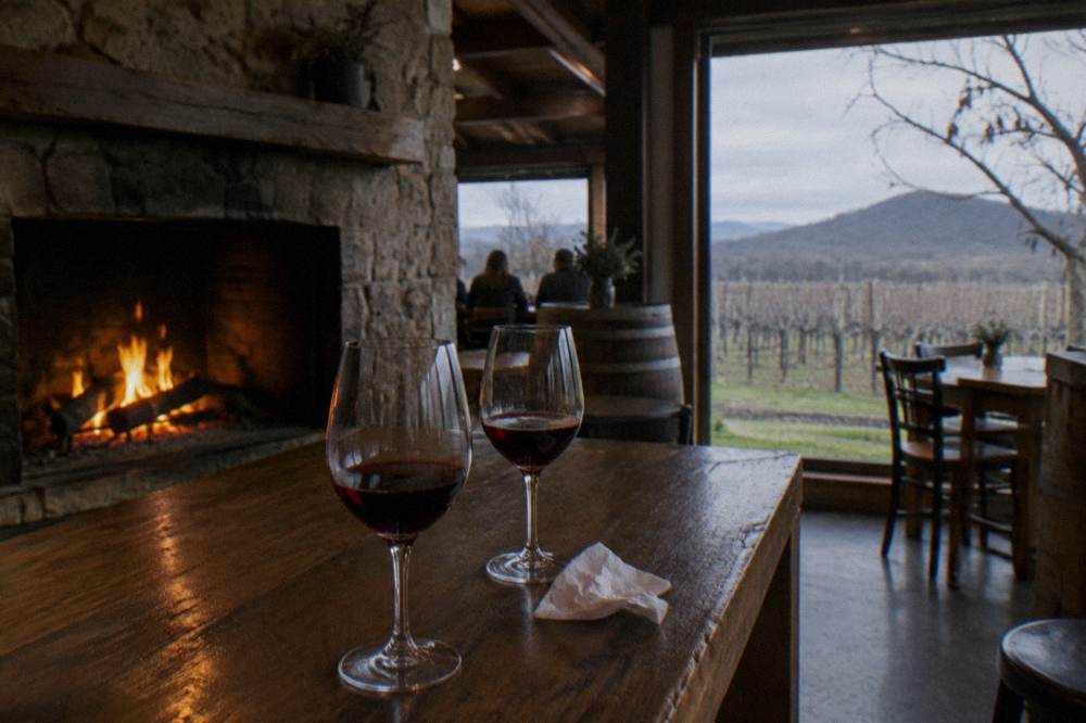 A rustic cellar door tasting room with a wood fire and glasses of red wine on a timber bar
