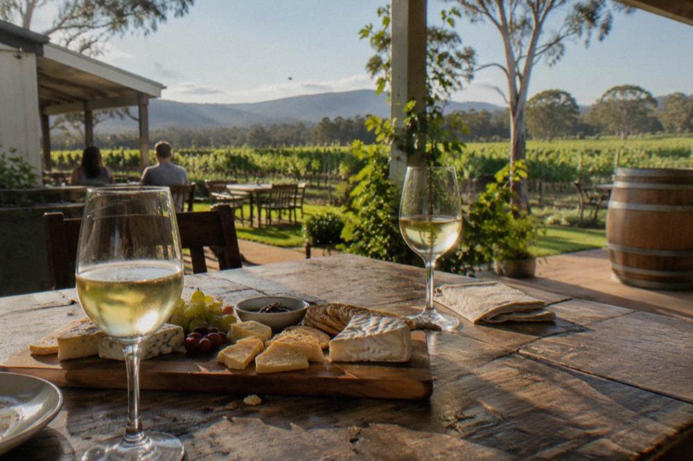 A wooden board of cheese wedges and crackers beside two glasses of white wine on a cellar door table.