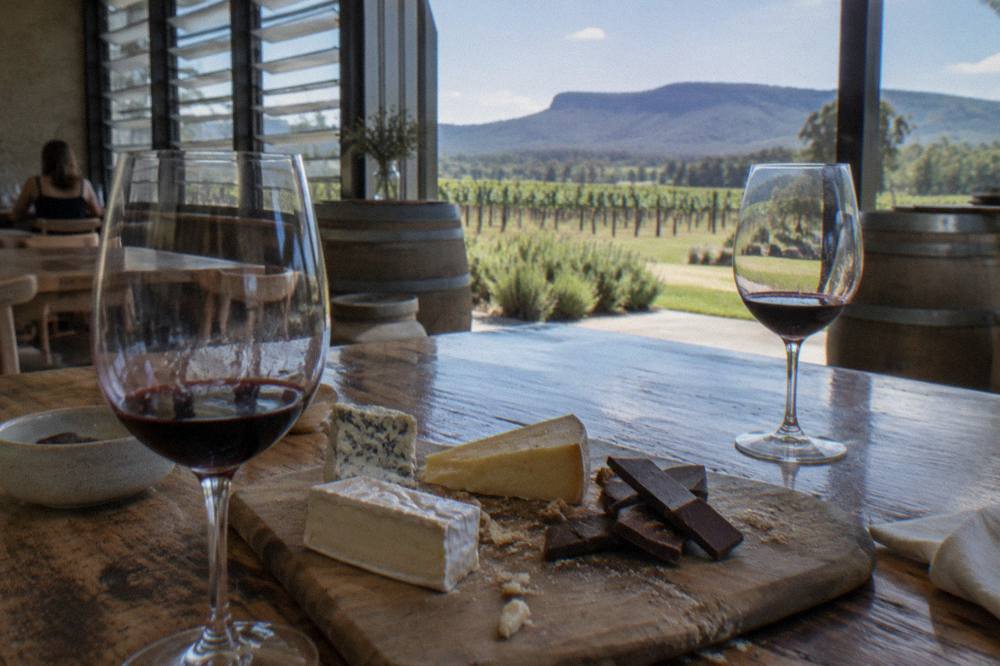 A tasting platter with cheese wedges, chocolate pieces and two glasses of red wine on a timber table