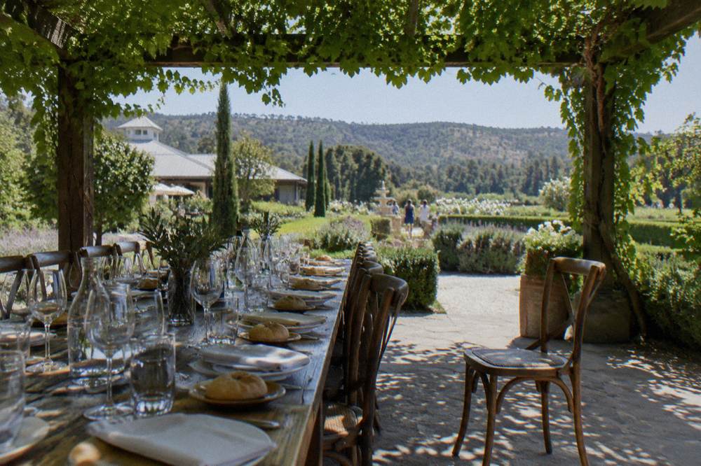 A long luncheon table set with wine glasses and bread rolls under a pergola