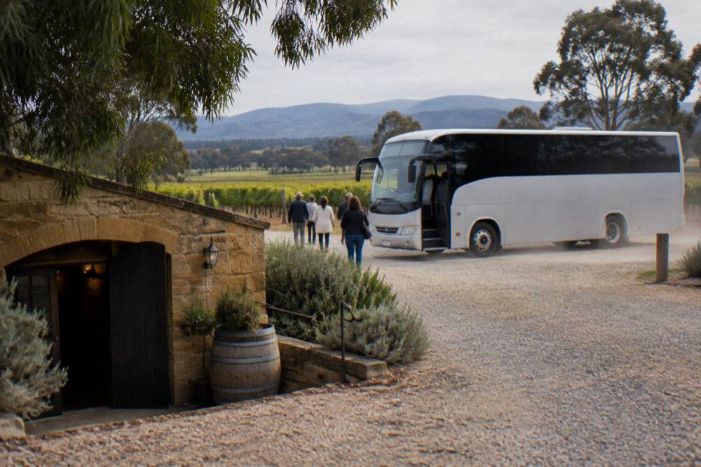 A tour bus waiting outside a low sandstone cellar door building among vines