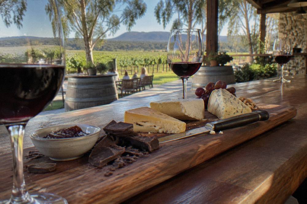 A wooden tasting board with cheese wedges, chocolate pieces and glasses of red wine on a cellar bar