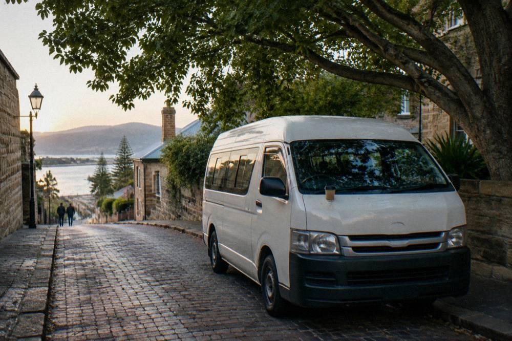 A small tour van parked on a gravel driveway beside rows of vineyard vines