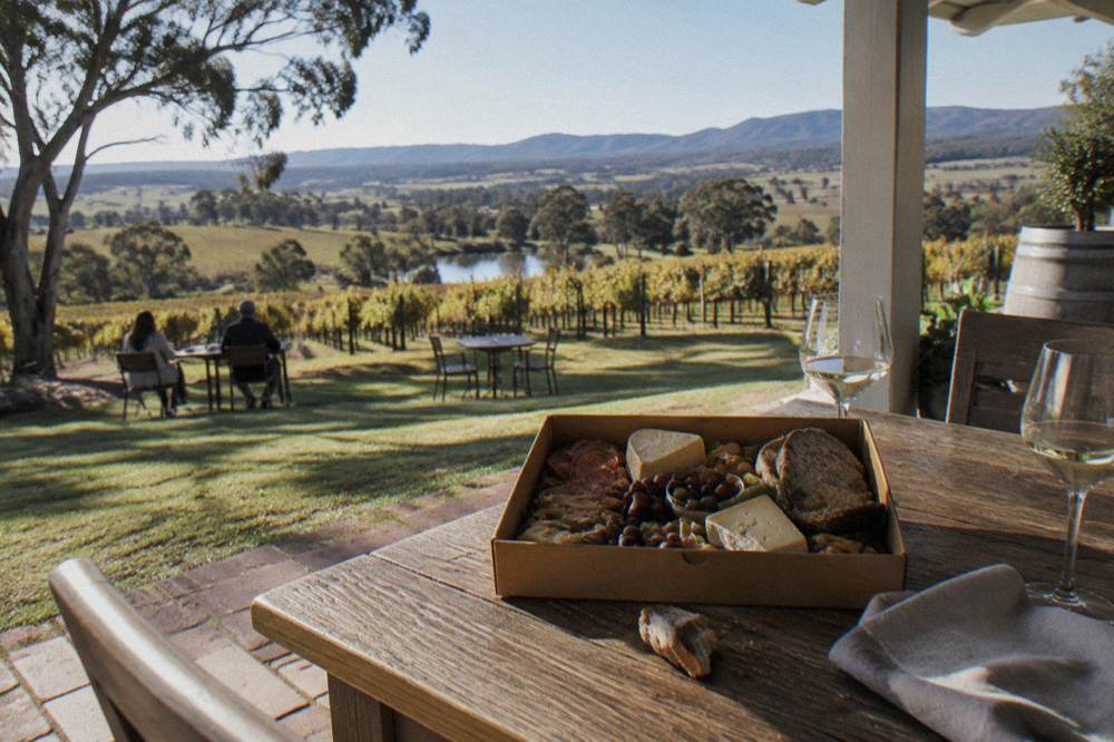 A grazing box of cheese, cured meat and bread on a wooden table overlooking vineyards