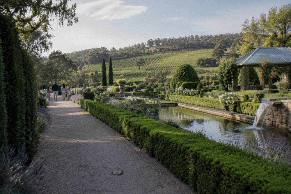 Formal garden beds and hedged paths with a vineyard backdrop under an afternoon sky