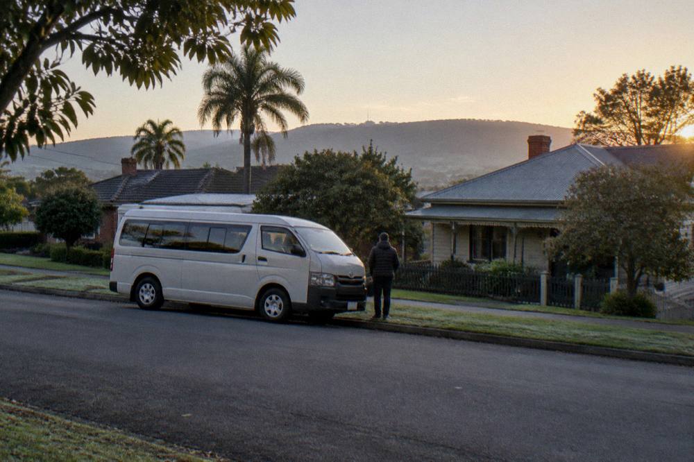 A small passenger van on a country road with vineyard rows stretching to distant hills at late afternoon