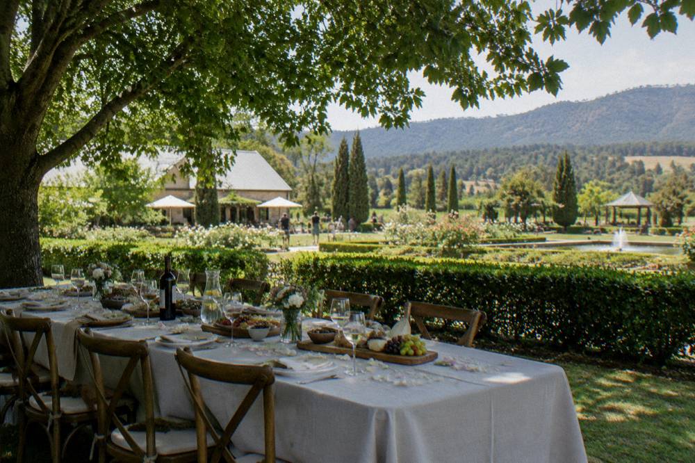 A long table set for lunch under trees beside a vineyard garden